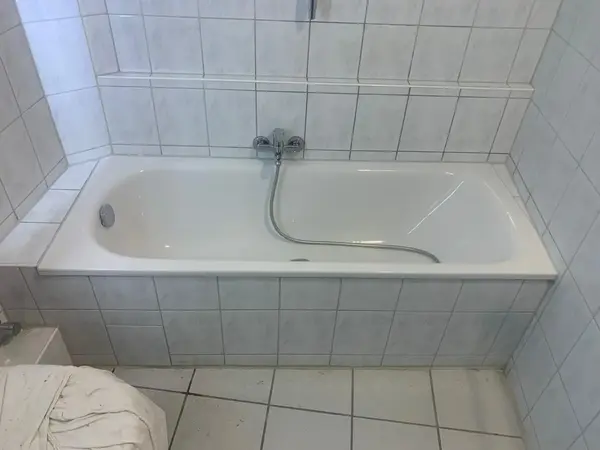 Weiße Badewanne mit Armatur und Duschschlauch, umgeben von hellen Fliesen im Badezimmer.