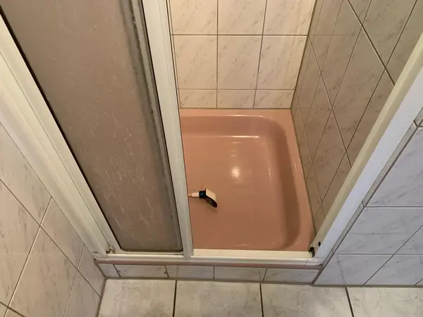 Duschkabine mit rosa Duschwanne, Fliesenwänden und Rasierer auf dem Boden.